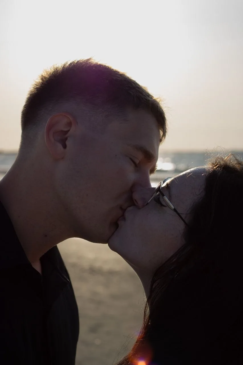 Close beach maternity moment with expecting parents kissing in warm backlight.